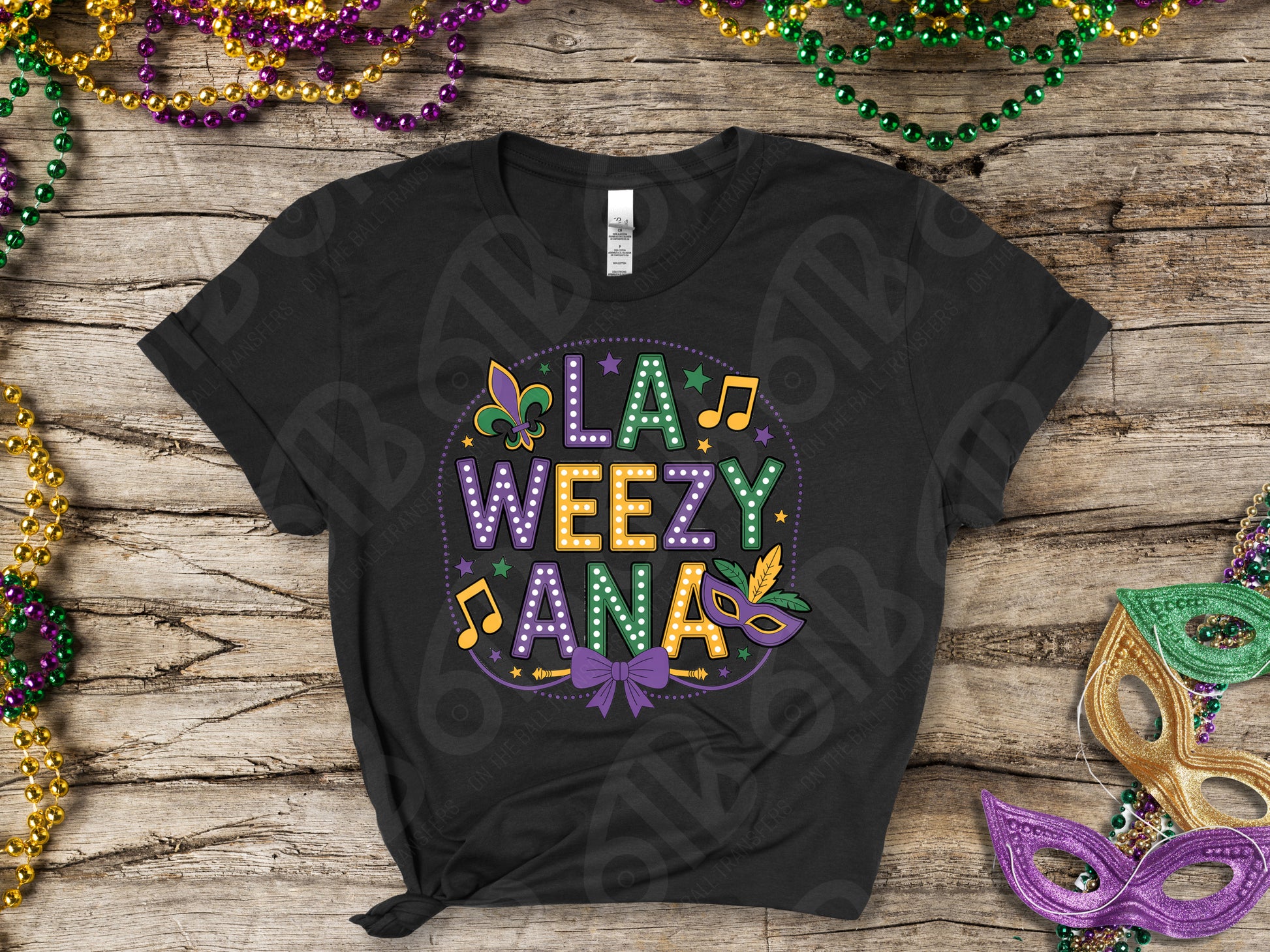 Black t-shirt with colorful 'LA WEEZY ANA' design on a wooden surface with Mardi Gras decorations.