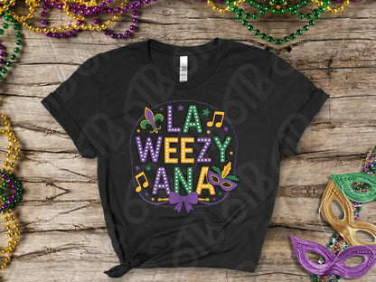 Black t-shirt with colorful 'LA WEEZY ANA' design on a wooden surface with Mardi Gras decorations.