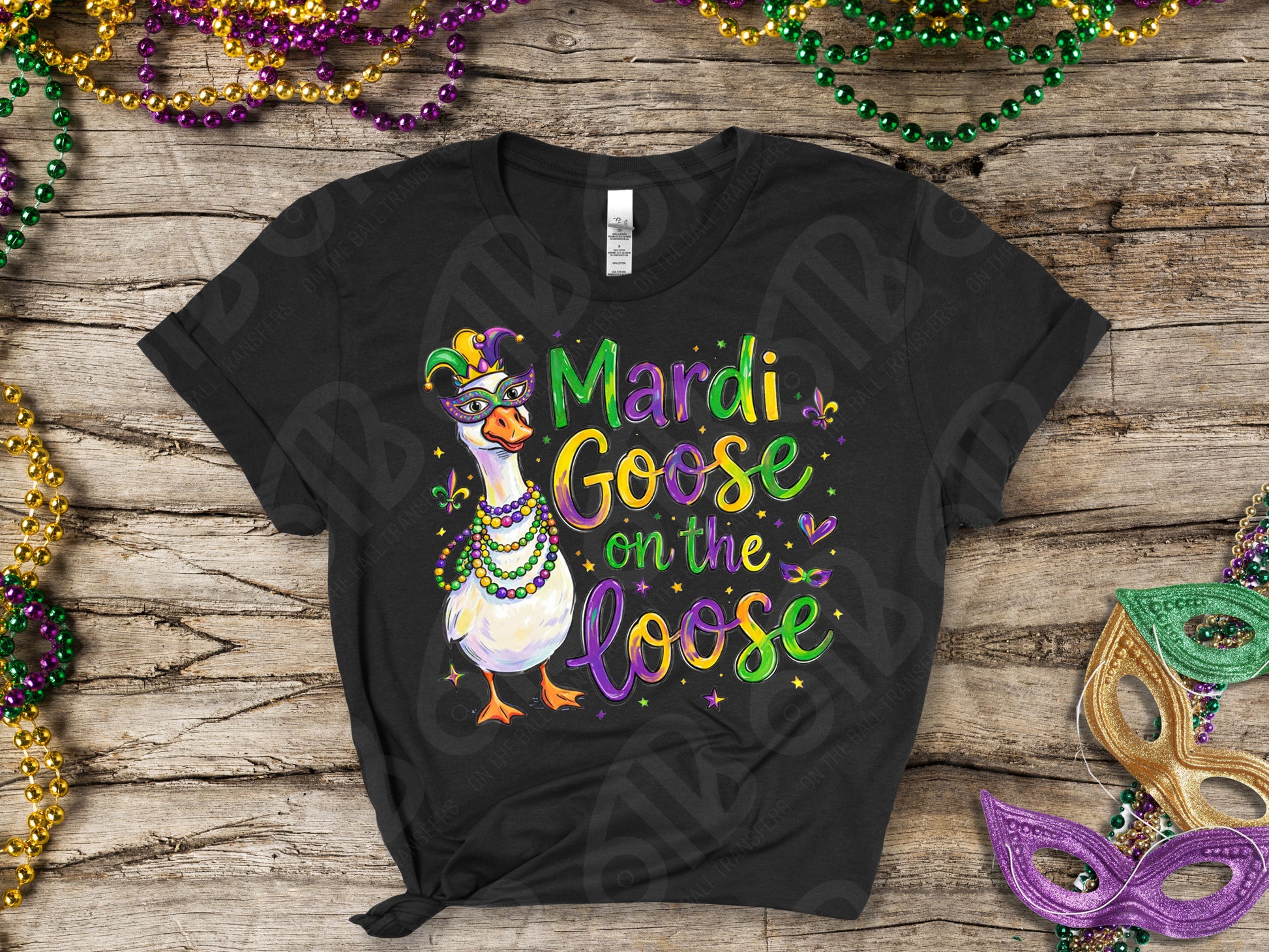 Black t-shirt with 'Mardi Goose on the Loose' design on a wooden surface with Mardi Gras decorations.