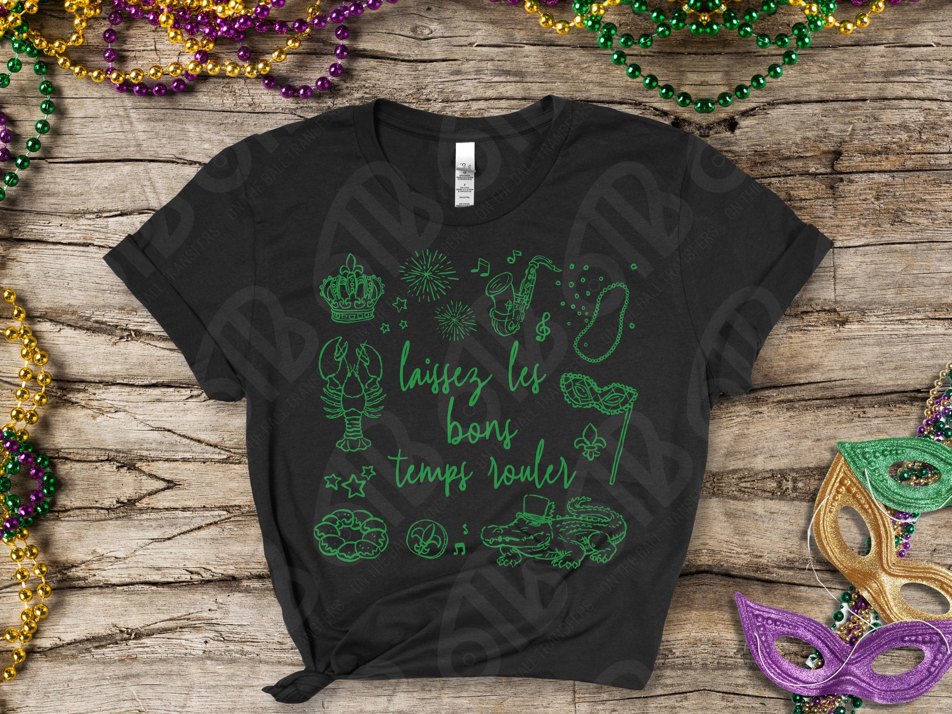 Black t-shirt with green graphics and text on a wooden surface with Mardi Gras decorations.