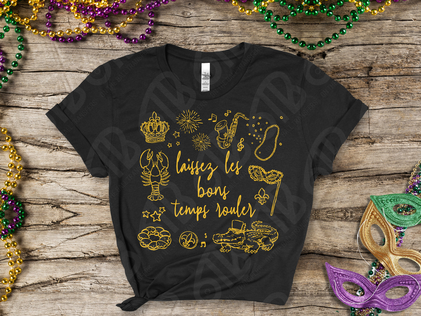 Black t-shirt with gold graphics and text on a wooden surface with Mardi Gras decorations.
