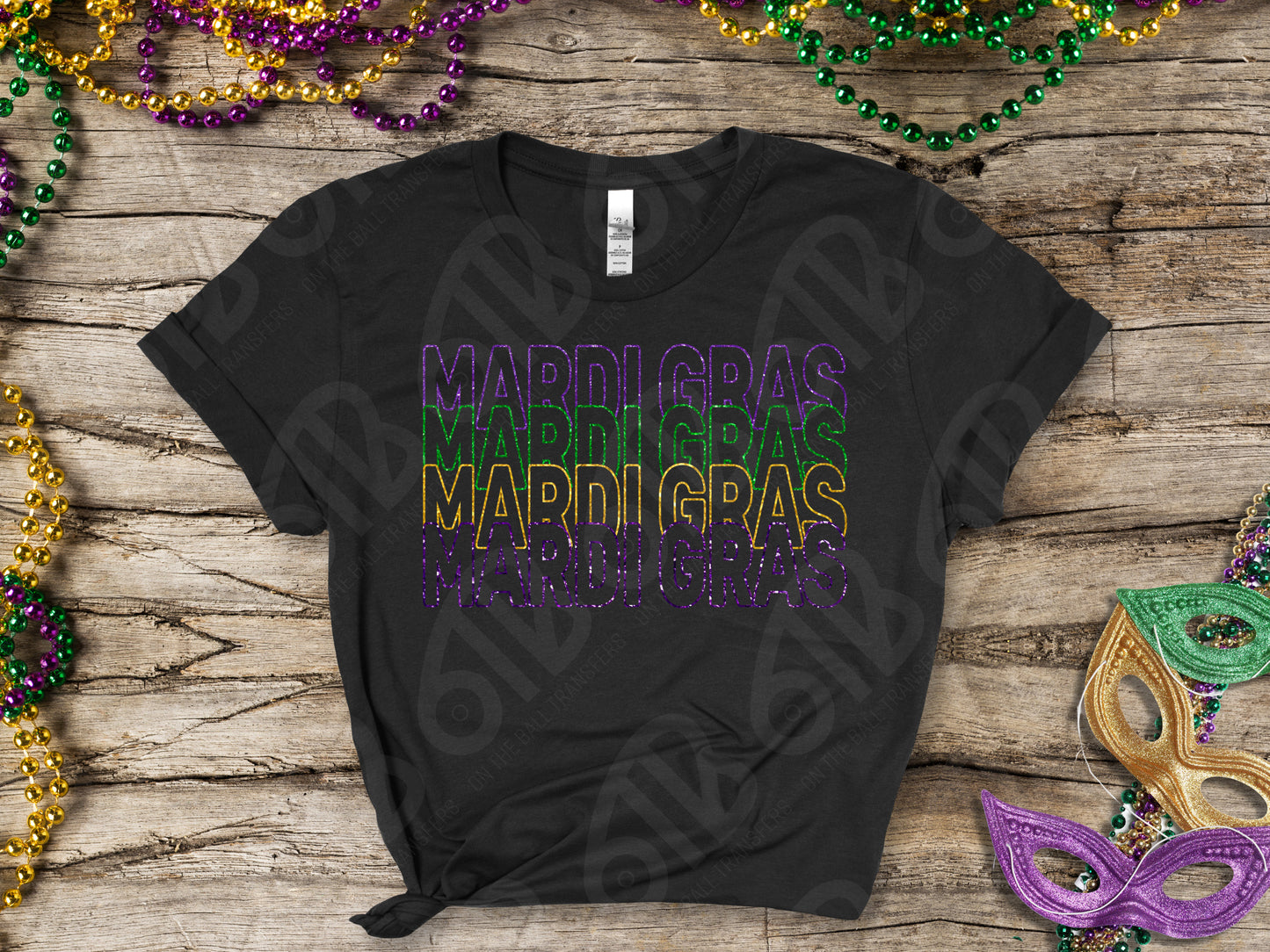 Black t-shirt with 'MARDI GRAS' text in purple, gold, and green on a wooden surface with Mardi Gras beads and a mask.